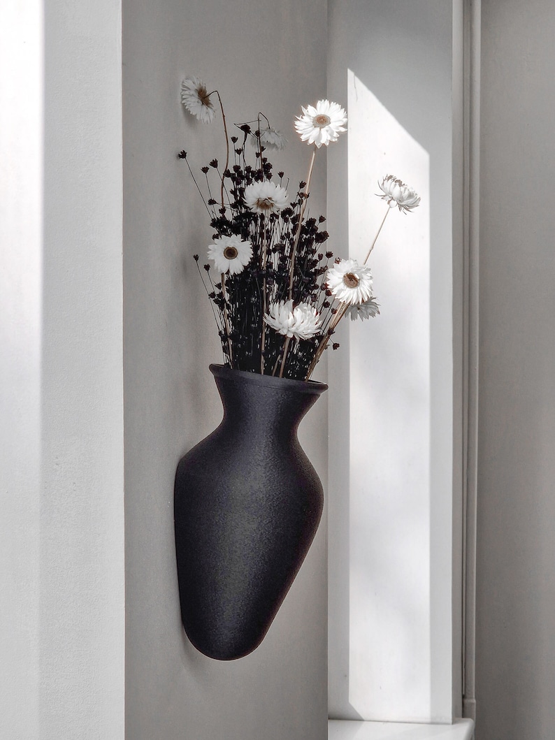 May include: A black ceramic vase with a textured finish, mounted on a white wall, holds a bouquet of dried white daisies.