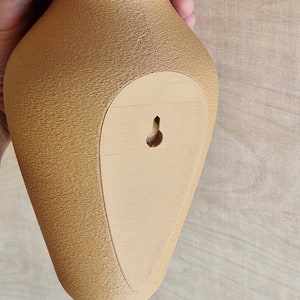 May include: A tan ceramic vase with a textured surface and a small hole on the back for hanging.