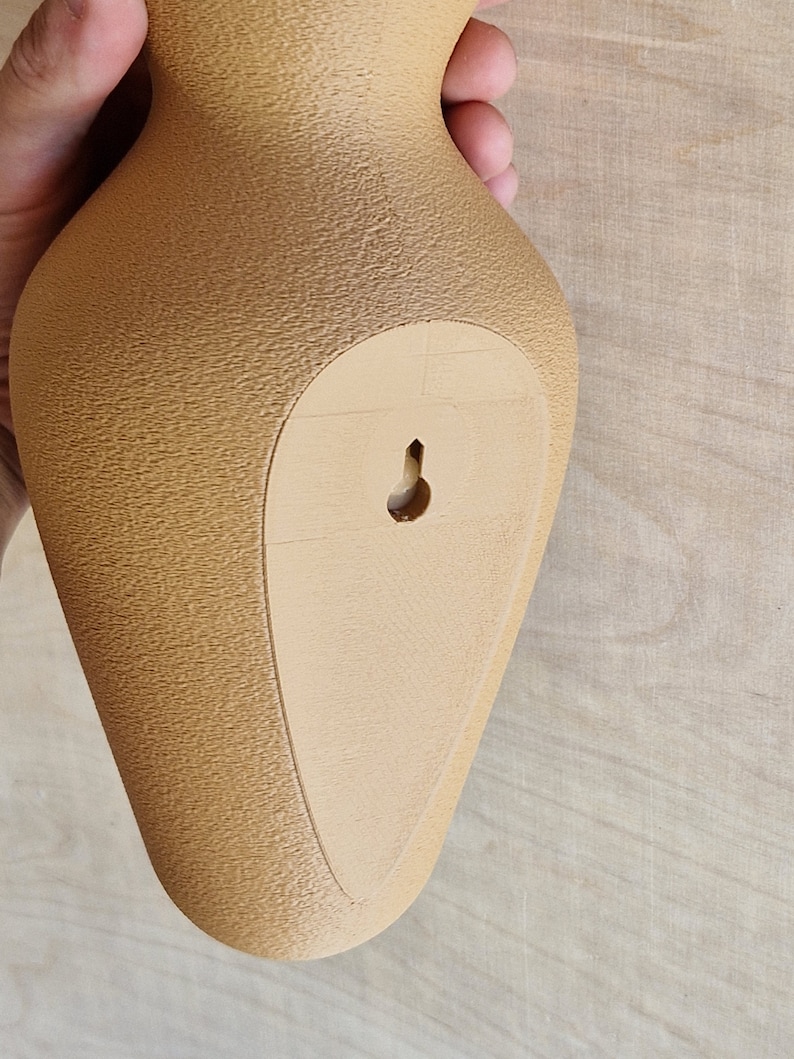 May include: A tan ceramic vase with a textured surface and a small hole on the back for hanging.