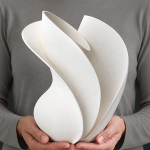 Puede incluir: Una escultura de cerámica blanca mate con formas fluidas, curvas y superpuestas que recuerdan a pétalos, creando una silueta dinámica en espiral. El objeto tiene una textura de superficie lisa y uniforme y una estructura hueca y abierta.