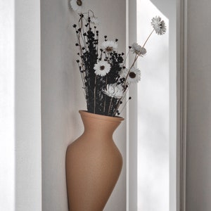 May include: A tan ceramic vase with a textured surface, mounted on a white wall, holds a bouquet of dried white flowers with black stems.