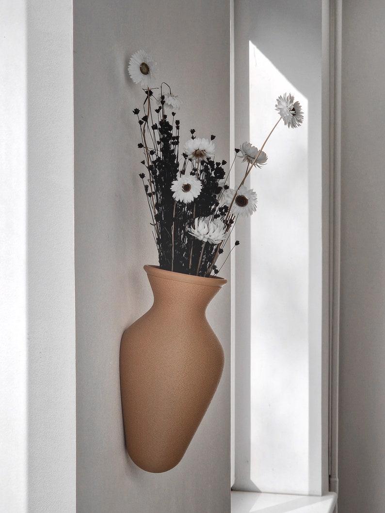 May include: A tan ceramic vase with a textured surface, mounted on a white wall, holds a bouquet of dried white flowers with black stems.