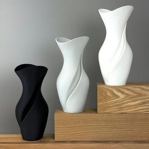 Puede incluir: Tres jarrones modernos en un estudio. Un jarrón es negro y los otros dos son blancos. Cada jarrón tiene un diseño único y fluido. Los jarrones se exhiben sobre una superficie de madera sobre un fondo gris. Los jarrones son decorativos.