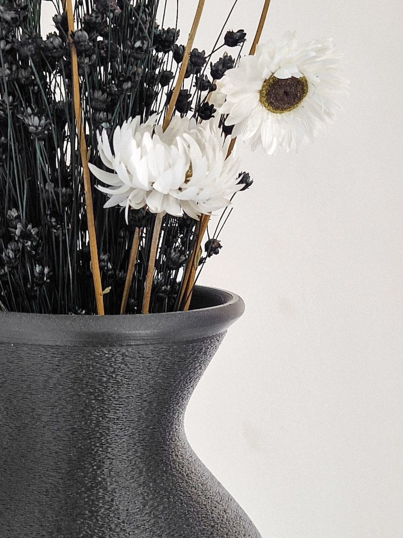 May include: A black ceramic vase with a textured surface holds a bouquet of dried white flowers and black dried grasses.
