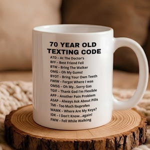 May include: A white ceramic mug featuring black printed text titled 70 YEAR OLD TEXTING CODE, followed by a list of humorous acronyms and their meanings, including ATD, BFF, BTW, OMG, BYOT, FWIW, OMSG, TGIF, APP, ASAP, TMI, WAMK, IDK, and FWW.