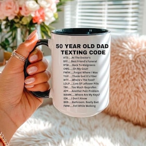 May include: A white ceramic mug with a black rim and handle, featuring the text "50 YEAR OLD DAD TEXTING CODE" and a list of acronyms. The mug is held by a person with manicured nails, with a blurred floral background.