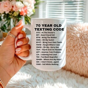 May include: White ceramic mug with the text "70 YEAR OLD TEXTING CODE" and a list of acronyms and their meanings. The mug is held by a person with manicured nails, with a blurred floral background.