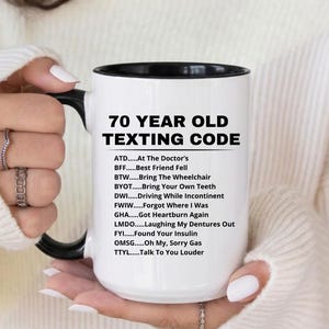 May include: A white ceramic mug with a black interior and handle, featuring the text "70 YEAR OLD TEXTING CODE" and a list of acronyms with their meanings. The mug is held by a person.