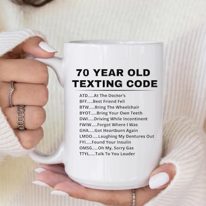 May include: A white ceramic mug with the text "70 YEAR OLD TEXTING CODE" in black. The mug lists common acronyms and phrases associated with aging, such as "At The Doctor's" and "Forgot Where I Was."