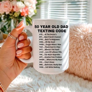 May include: White ceramic mug with the text "50 YEAR OLD DAD TEXTING CODE" in black. The mug lists common texting abbreviations. The mug is held by a person with manicured nails, with a blurred background of flowers and a pillow.
