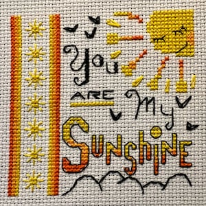 Puede incluir: Bordado de punto de cruz con hilo amarillo y naranja sobre tela blanca. El diseño presenta un sol con rayos, un corazón y el texto "You are my sunshine".