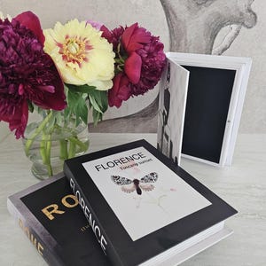 Könnte beinhalten: Ein Stillleben mit zwei dekorativen Büchern, eines mit "ROME" in Goldbuchstaben und das andere mit "FLORENCE Tuscany sunset". Eine Vase mit Pfingstrosen in Burgunderrot und Gelb steht neben einem weißen Rahmen.