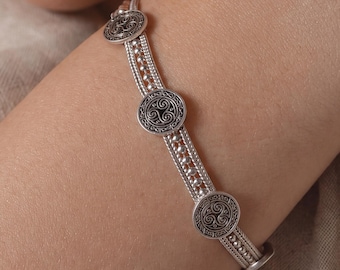 May include: A silver-toned metal bracelet featuring three circular medallions with intricate spiral patterns. The band is constructed from two parallel beaded chains that connect the medallions, creating a textured and detailed design.