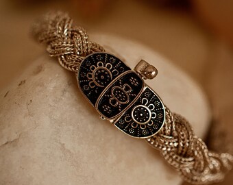 May include: A braided metallic bracelet featuring a central oval-shaped clasp. The clasp is black with intricate gold-toned floral and geometric patterns. The bracelet band consists of a woven, textured metallic chain that connects to the clasp.