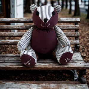 May include: A plush teddy bear with a burgundy body and head, and striped arms, legs, and ears. The bear is sitting on a wooden bench. The bear's feet are embroidered with the names "Graciela" and "Abel".