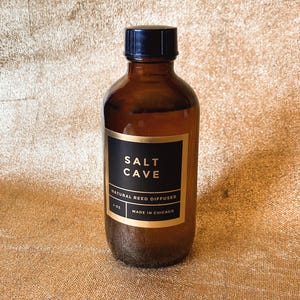 May include: An amber glass bottle with a black screw-top lid, featuring a black label with gold trim. The label reads SALT CAVE, NATURAL REED DIFFUSER, 4 OZ, and MADE IN CHICAGO in white and gold lettering.