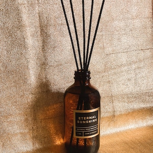 May include: An amber glass reed diffuser bottle with five black reeds inserted. The rectangular label features the text ETERNAL SUNSHINE, NATURAL REED DIFFUSER, 8 OZ, and MADE IN CHICAGO in white lettering against a black background.
