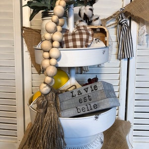 Op de afbeelding: Een witte etagère met een houten kralenslinger, een bruin en wit geruit stoffen hart, een houten bord met de tekst "La vie est belle" en een banner van jute.