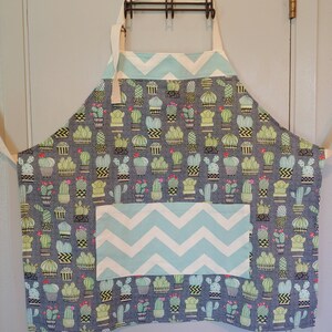 May include: A blue and white chevron patterned kitchen apron with a large pocket. The apron is covered in a repeating pattern of potted cacti in various shades of green.