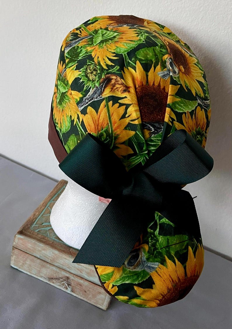 May include: A green, brown, and yellow surgical scrub hat with a sunflower print and a dark green bow.