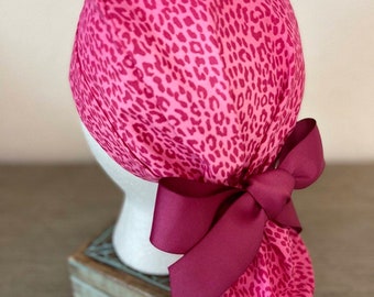 May include: A pink scrub cap featuring a leopard print design. The cap is tied with a burgundy bow at the back. The cap is displayed on a white mannequin head.