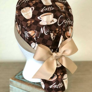 May include: A brown and tan scrub hat with a coffee theme. The hat has a brown and white checkered pattern with coffee cups, coffee beans, and the words "Cappuccino", "Latte", "Mocha", "Espresso", and "Coffee". The hat has a tan bow tie.