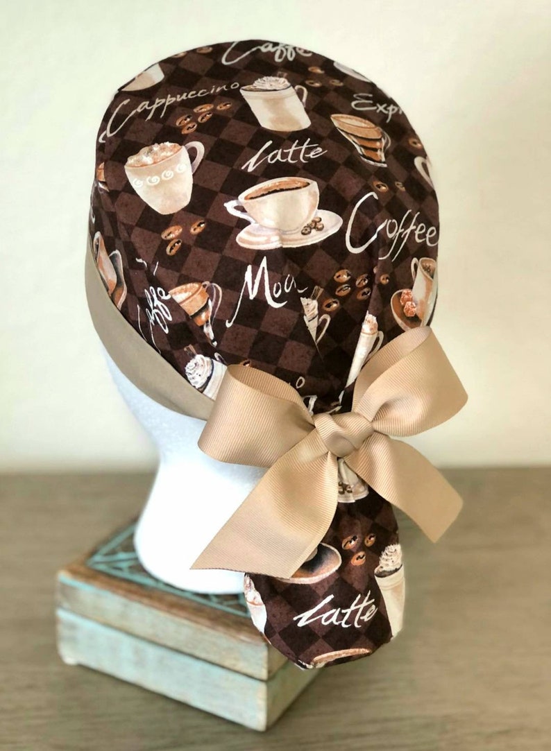 May include: A brown and tan scrub hat with a coffee theme. The hat has a brown and white checkered pattern with coffee cups, coffee beans, and the words "Cappuccino", "Latte", "Mocha", "Espresso", and "Coffee". The hat has a tan bow tie.