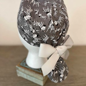 May include: Gray scrub cap with a white zebra print and a light gray bow. The cap is tied in the back with a large bow. The cap is on a white mannequin head.