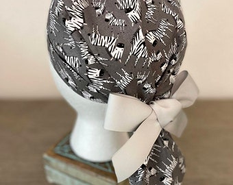 May include: Gray scrub cap with a white zebra print and a light gray bow. The cap is tied in the back with a large bow. The cap is on a white mannequin head.