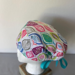May include: A colorful scrub cap featuring a leaf pattern in shades of pink, green, blue, and purple on a white background. The cap is tied with a turquoise ribbon. The cap is displayed on a white mannequin head.