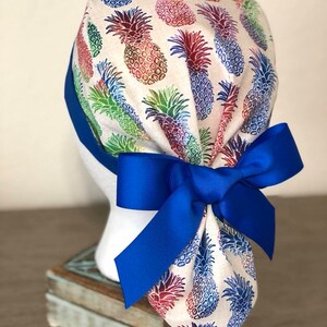 May include: A white scrub hat with a colorful pineapple print and a bright blue ribbon tied in a bow at the back.