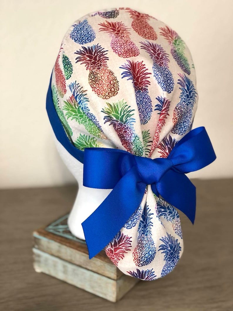 May include: A white scrub hat with a colorful pineapple print and a bright blue ribbon tied in a bow at the back.