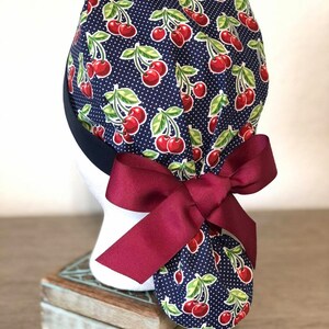 May include: A navy blue surgical scrub hat with a cherry print and white polka dots. The hat has a burgundy ribbon tied in a bow at the back.
