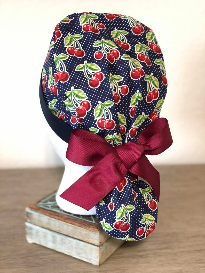 May include: A navy blue surgical scrub hat with a cherry print and white polka dots. The hat has a burgundy ribbon tied in a bow at the back.