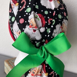 Op de afbeelding: Een zwarte operatiemuts met een herhalend patroon van vintage kerstmangezichten, rood-witte zuurstokken en groen-witte pepermuntjes op een achtergrond met witte stippen, afgewerkt met een felgroene strik van grosgrain lint.