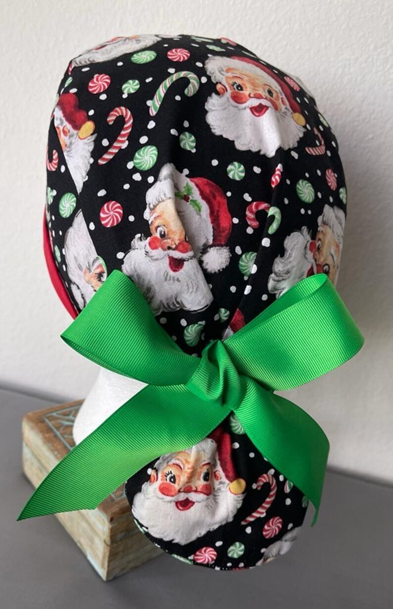 Op de afbeelding: Een zwarte operatiemuts met een herhalend patroon van vintage kerstmangezichten, rood-witte zuurstokken en groen-witte pepermuntjes op een achtergrond met witte stippen, afgewerkt met een felgroene strik van grosgrain lint.