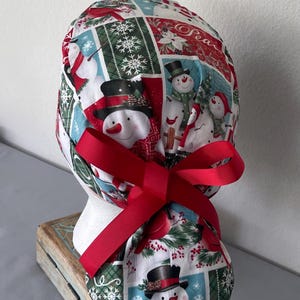 Op de afbeelding: Een operatiemuts met een feestelijke kerstprint van sneeuwpoppen, sneeuwvlokken en rode kardinalen in een rasterpatroon. De stof is aan de achterkant gestrikt met een effen rood grosgrain lint, wat zorgt voor een geplooide en verstelbare pasvorm.