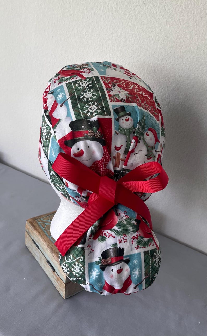 Op de afbeelding: Een operatiemuts met een feestelijke kerstprint van sneeuwpoppen, sneeuwvlokken en rode kardinalen in een rasterpatroon. De stof is aan de achterkant gestrikt met een effen rood grosgrain lint, wat zorgt voor een geplooide en verstelbare pasvorm.