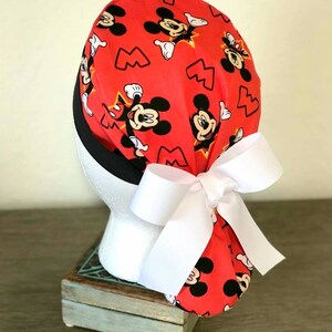 May include: Red scrub hat with a white bow and a Mickey Mouse print. The hat has black trim and a white bow.