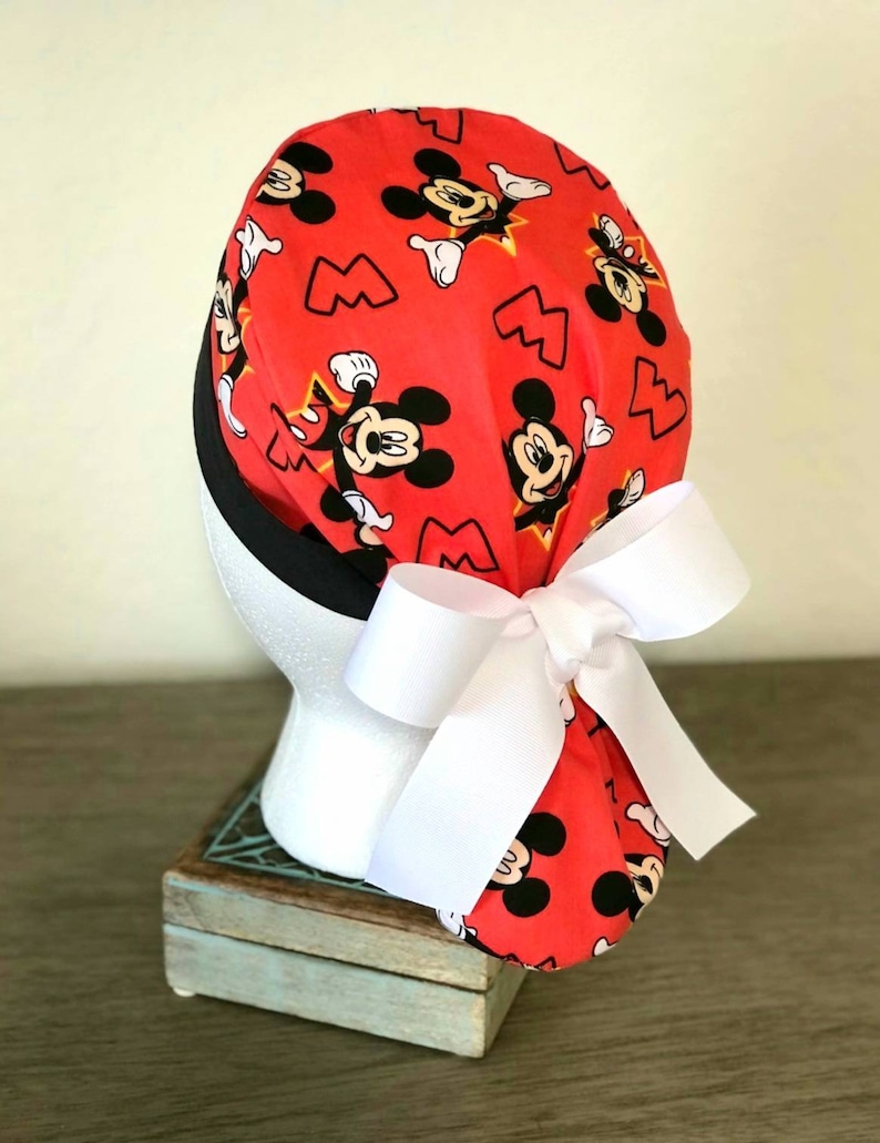 May include: Red scrub hat with a white bow and a Mickey Mouse print. The hat has black trim and a white bow.