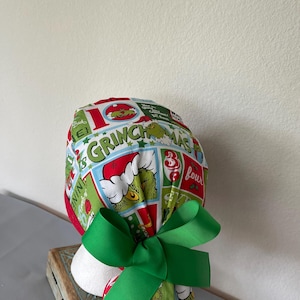 Op de afbeelding: Een operatiemuts met een kleurrijke Grinch-print in rode, groene en witte vlakken. De stof toont diverse illustraties van de Grinch, kerstafbeeldingen en de tekst Grinchmas. Een effen groen lint is aan de achterkant tot een strik gebonden.