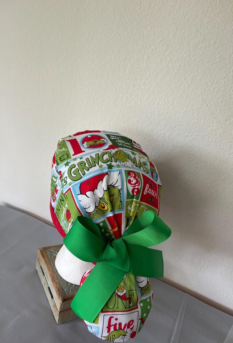 Op de afbeelding: Een operatiemuts met een kleurrijke Grinch-print in rode, groene en witte vlakken. De stof toont diverse illustraties van de Grinch, kerstafbeeldingen en de tekst Grinchmas. Een effen groen lint is aan de achterkant tot een strik gebonden.