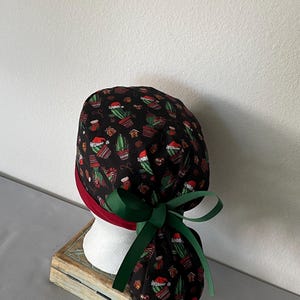 Op de afbeelding: Een zwarte operatiemuts met een herhalend patroon van groene cactussen met rode kerstmutsen, kleine peperkoekhuisjes en rode sokken. De muts heeft een effen donkerrode band aan de onderkant en wordt achteraan vastgemaakt met een donkergroen lint.