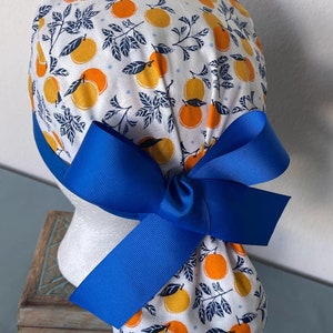 May include: A white scrub hat with a blue ribbon tied in a bow. The hat has a pattern of orange citrus fruit and dark blue leaves on a white background.