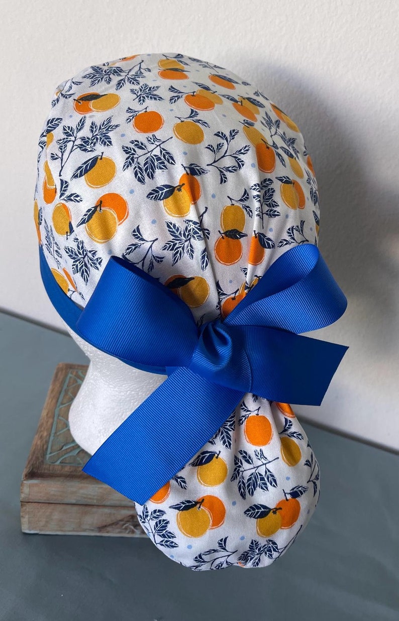 May include: A white scrub hat with a blue ribbon tied in a bow. The hat has a pattern of orange citrus fruit and dark blue leaves on a white background.