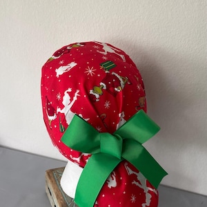 Op de afbeelding: Een rode operatiemuts met een herhalend patroon van groene Grinch-figuren, witte silhouetten en witte sterren. De muts is voorzien van een grote, effen groene grosgrain lintstrik aan de achterkant, wat zorgt voor een paardenstaartstijl.