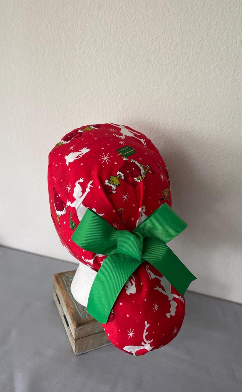 Op de afbeelding: Een rode operatiemuts met een herhalend patroon van groene Grinch-figuren, witte silhouetten en witte sterren. De muts is voorzien van een grote, effen groene grosgrain lintstrik aan de achterkant, wat zorgt voor een paardenstaartstijl.
