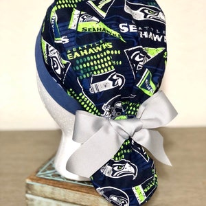 Op de afbeelding: Een marineblauwe operatiemuts met een herhalend patroon van Seattle Seahawks-logo's, tekst en wimpels in limoengroen, wit en donkerblauw. De muts heeft een effen marineblauwe band en een lichtgrijs lint dat achteraan tot een strik is geknoopt.