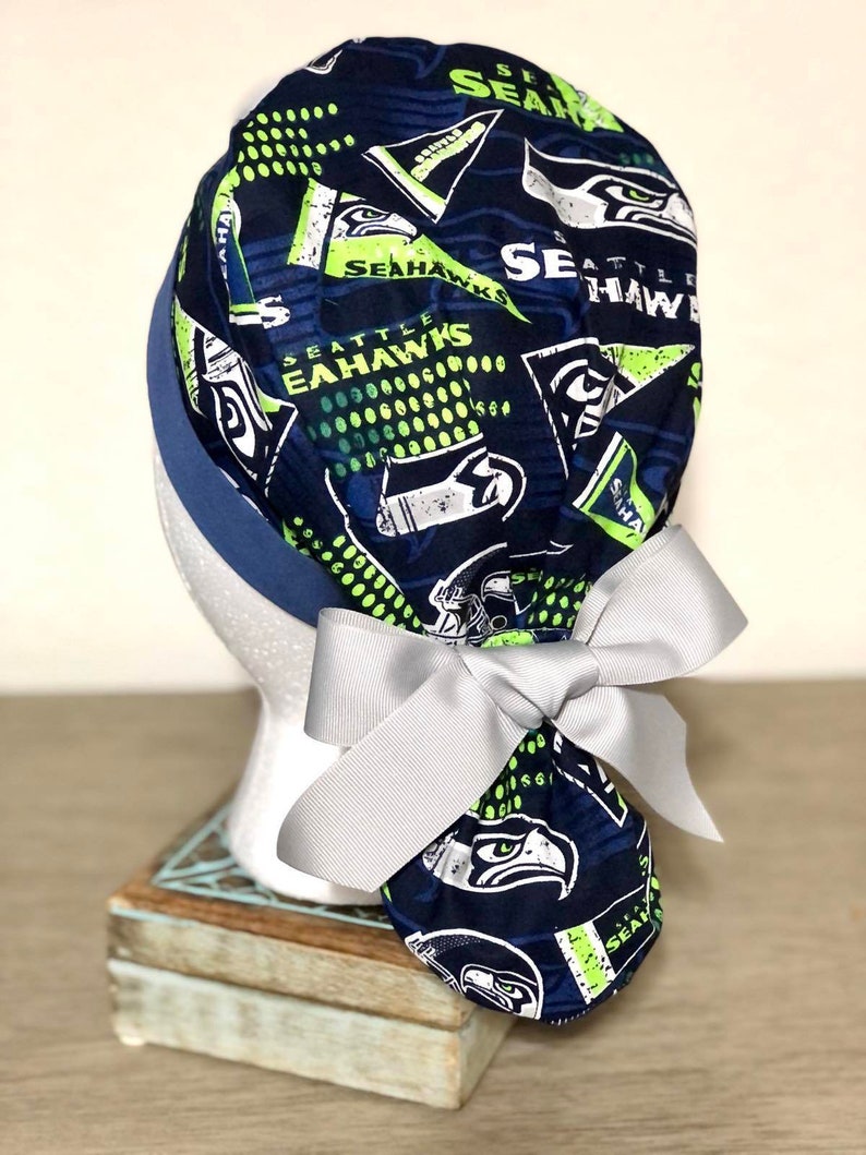 Op de afbeelding: Een marineblauwe operatiemuts met een herhalend patroon van Seattle Seahawks-logo's, tekst en wimpels in limoengroen, wit en donkerblauw. De muts heeft een effen marineblauwe band en een lichtgrijs lint dat achteraan tot een strik is geknoopt.