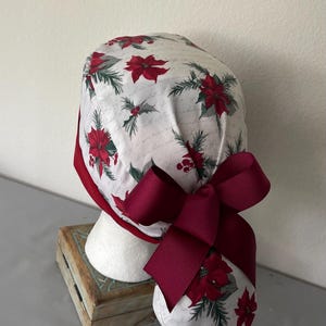 Op de afbeelding: Een operatiemuts met een witte achtergrond voorzien van een patroon van rode kerststerren, groene dennennaalden en vage cursieve tekst. De muts heeft een effen donkerrode rand aan de onderkant en een grote strik van donkerrood grosgrain lint.
